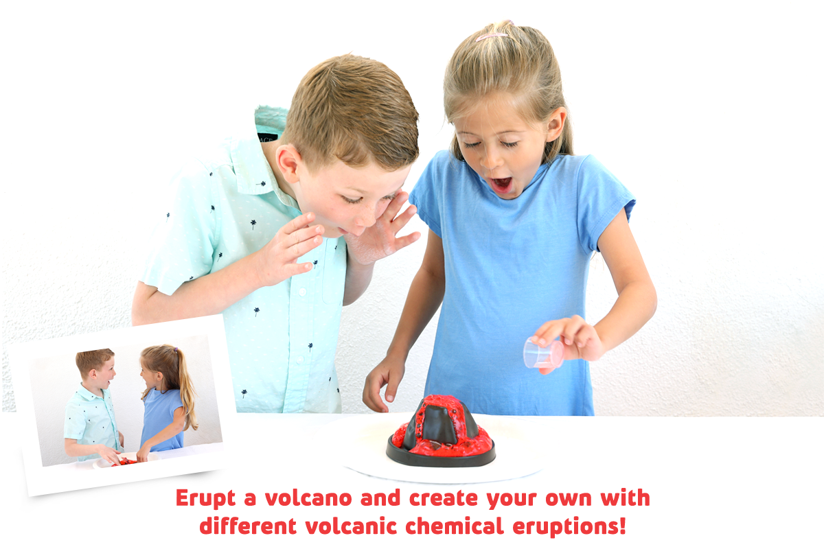 Two children experimenting with a red and black volcano eruption kit on a white table against a white wall. One child is covering their mouth as if emitting steam, and the other is holding a small clear container.